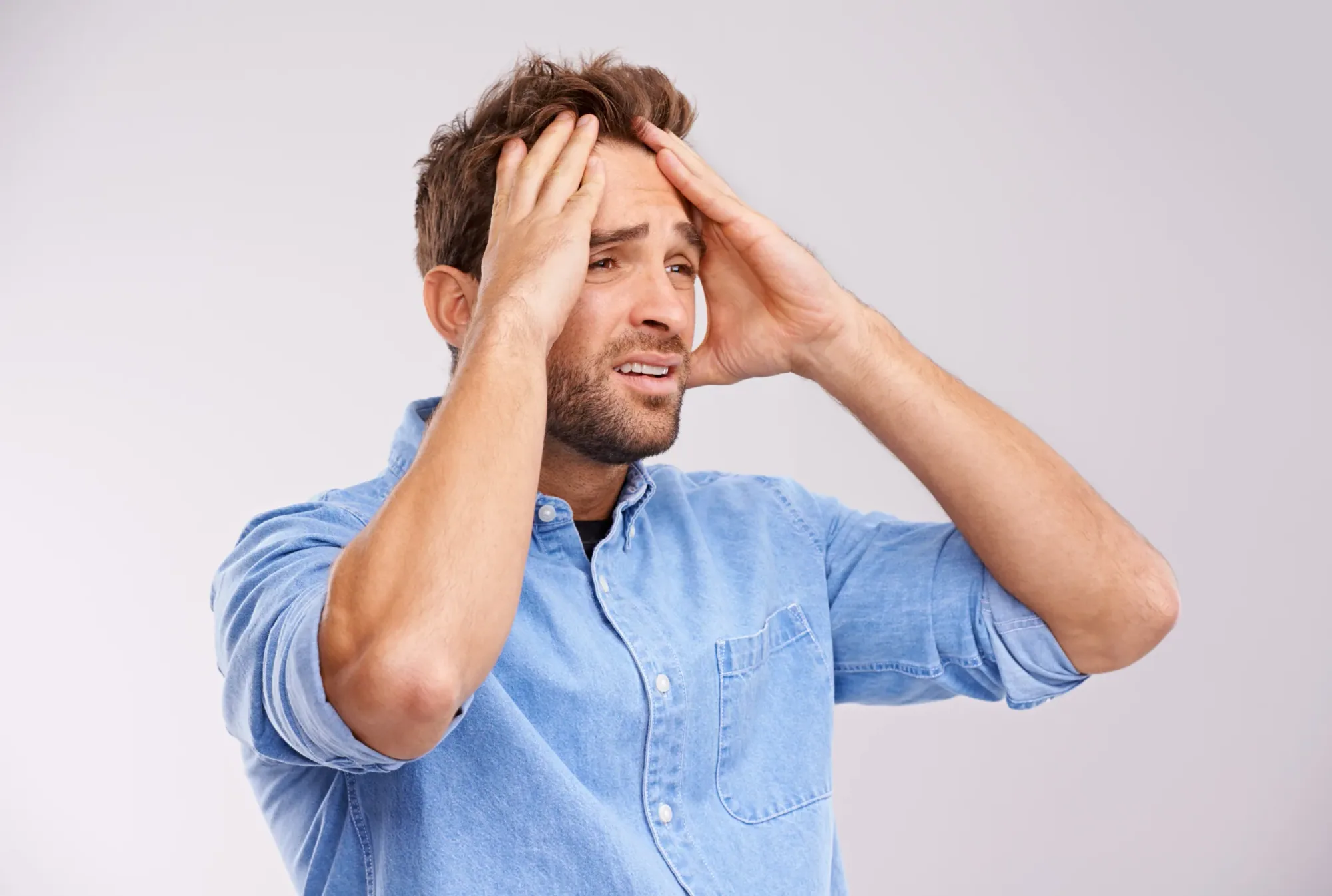 A man standing against a pale grey background with both hands raised to his head, appearing overwhelmed.